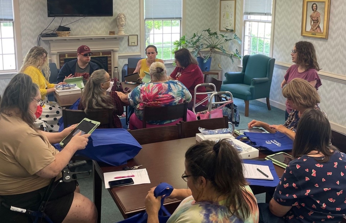 Image of windowed room with two large brown tables on blue-green carpet, and each table has 4-5 individuals of various ages working on iPads. A woman with red curly hair and a mauve shirt is talking to the entire class.